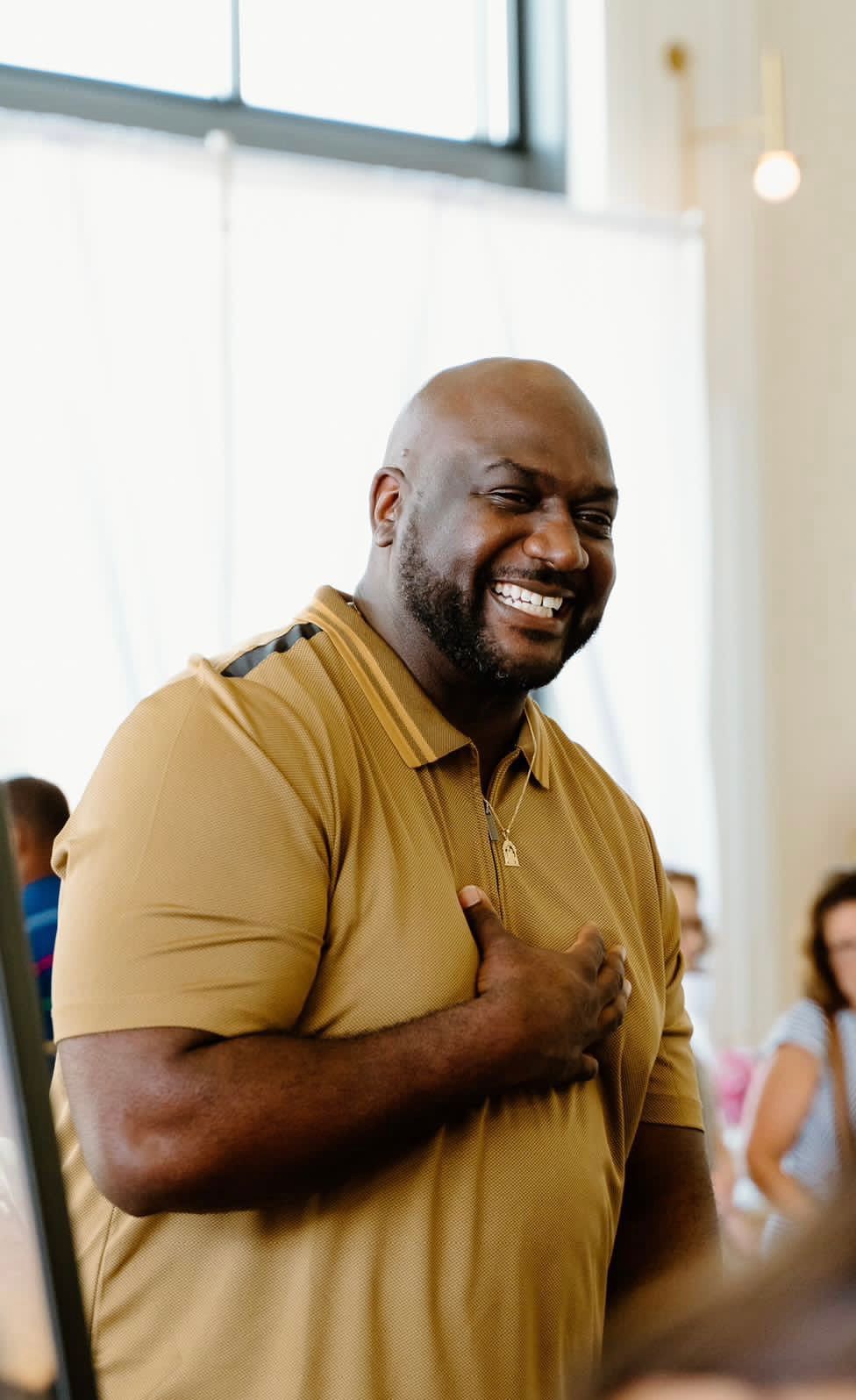 Smiling bald Black man in a tan polo shirt gesturing with his hand in a bright indoor setting.