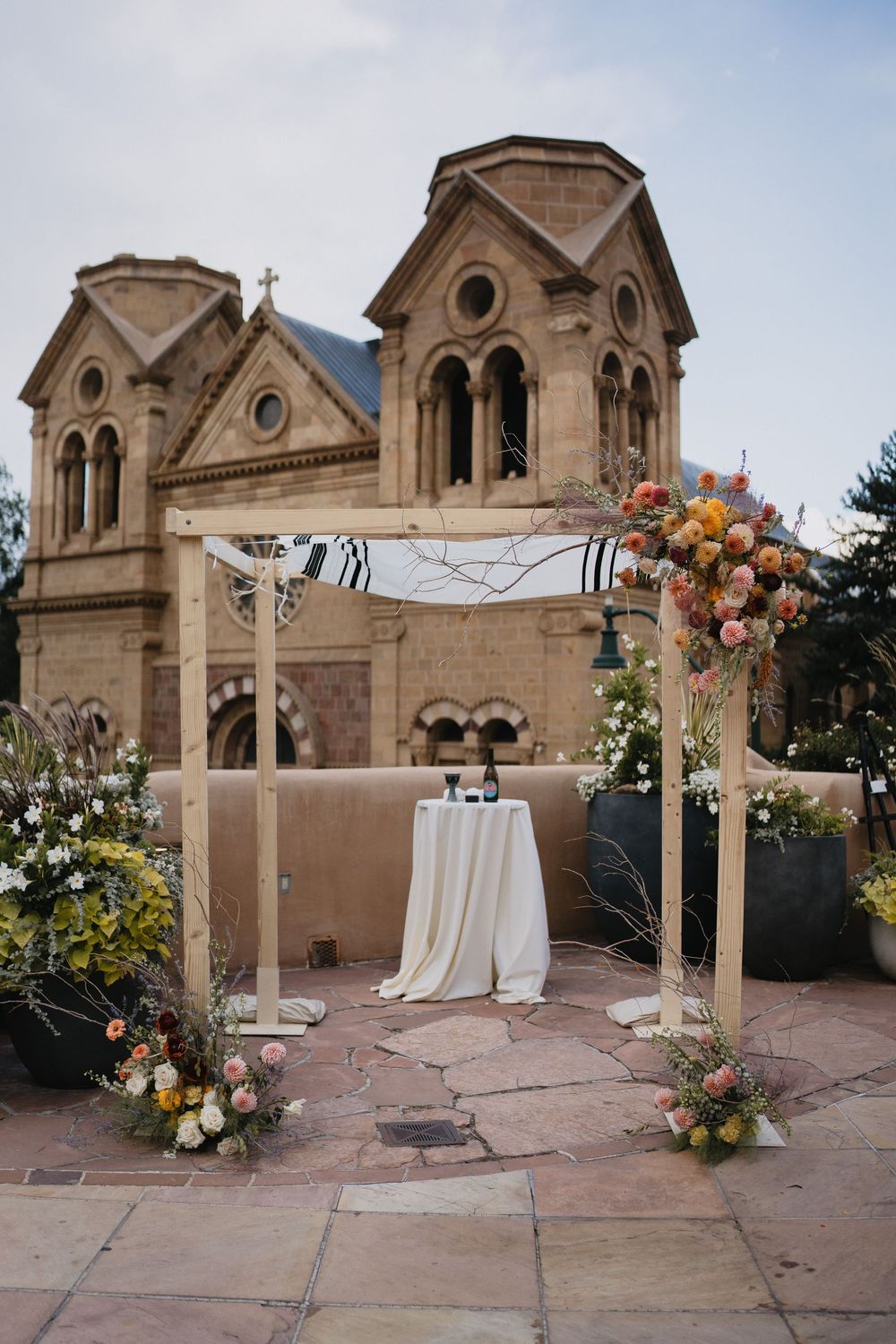 Outdoor wedding chuppah with colorful floral arrangements on stone patio, historic Cathedral Basilica of Saint Francis in background.