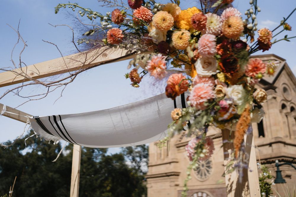 Outdoor wedding chuppah adorned with colorful dahlias in peach, orange, and pink tones, with a church visible in the background.