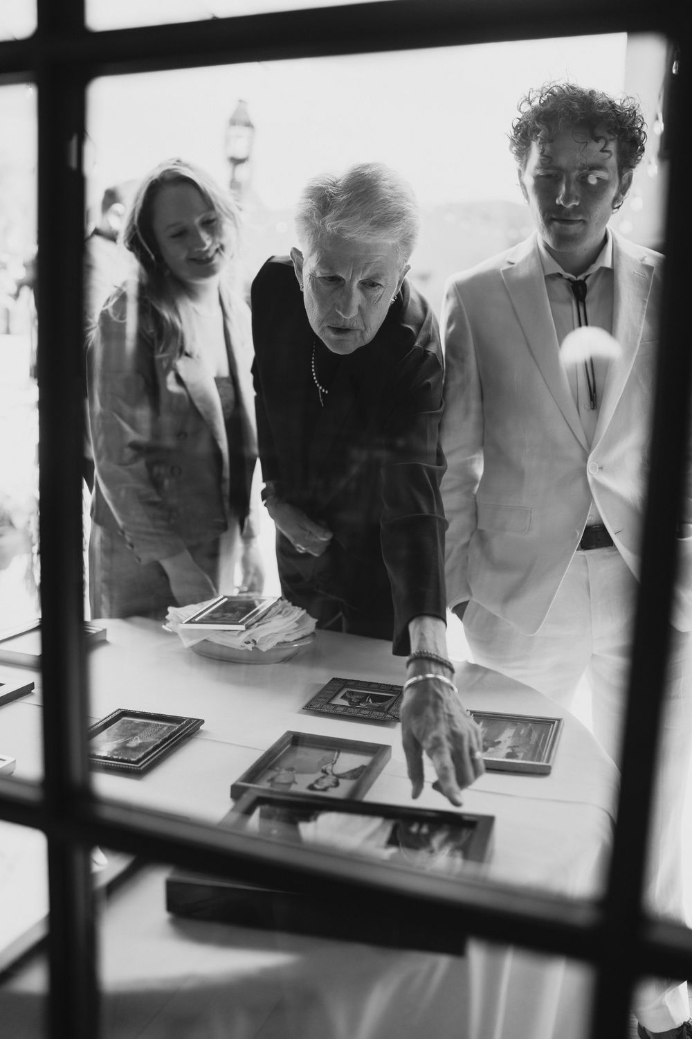 Black and white photo of three people examining framed photographs on a table indoors.