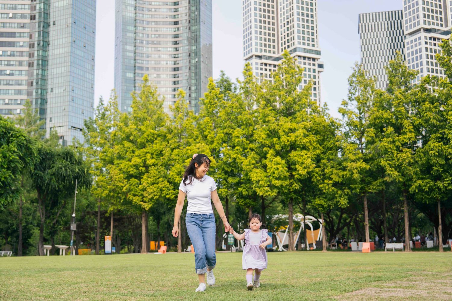 A mother and young daughter walk hand-in-hand through a lush urban park with tall skyscrapers in the background.
