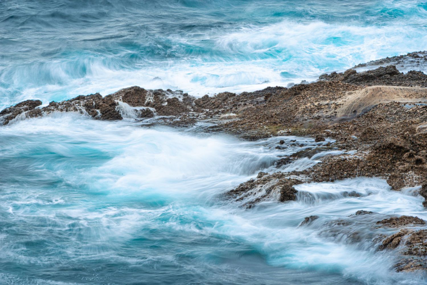 Turquoise ocean waves crashing over rocky coastline with white sea foam and blue water in a long exposure coastal landscape.