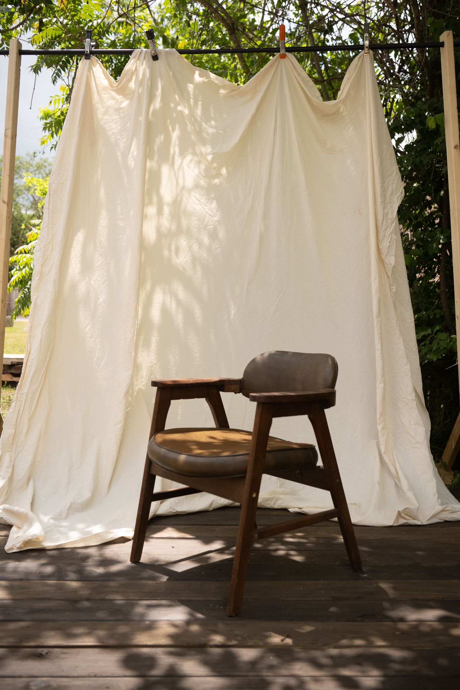 Vintage wooden armchair with leather cushion on a deck against white draped fabric backdrop outdoors.