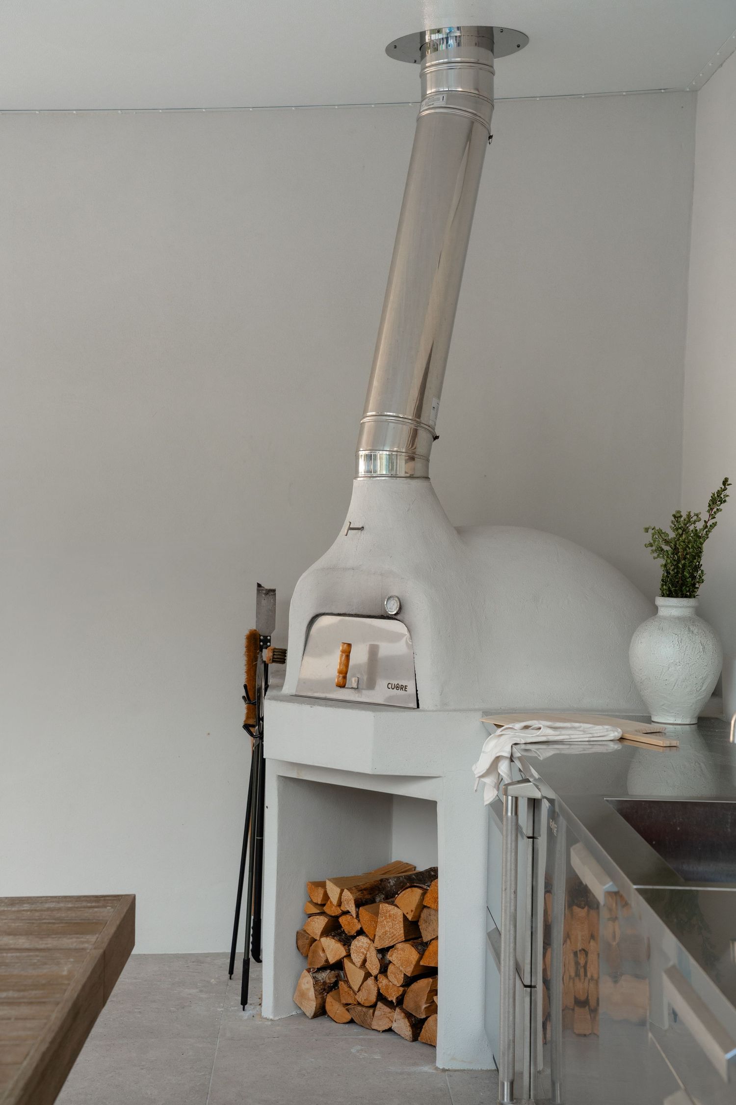 White wood-fired pizza oven with stainless steel chimney, firewood storage below, and fireplace tools in modern kitchen.