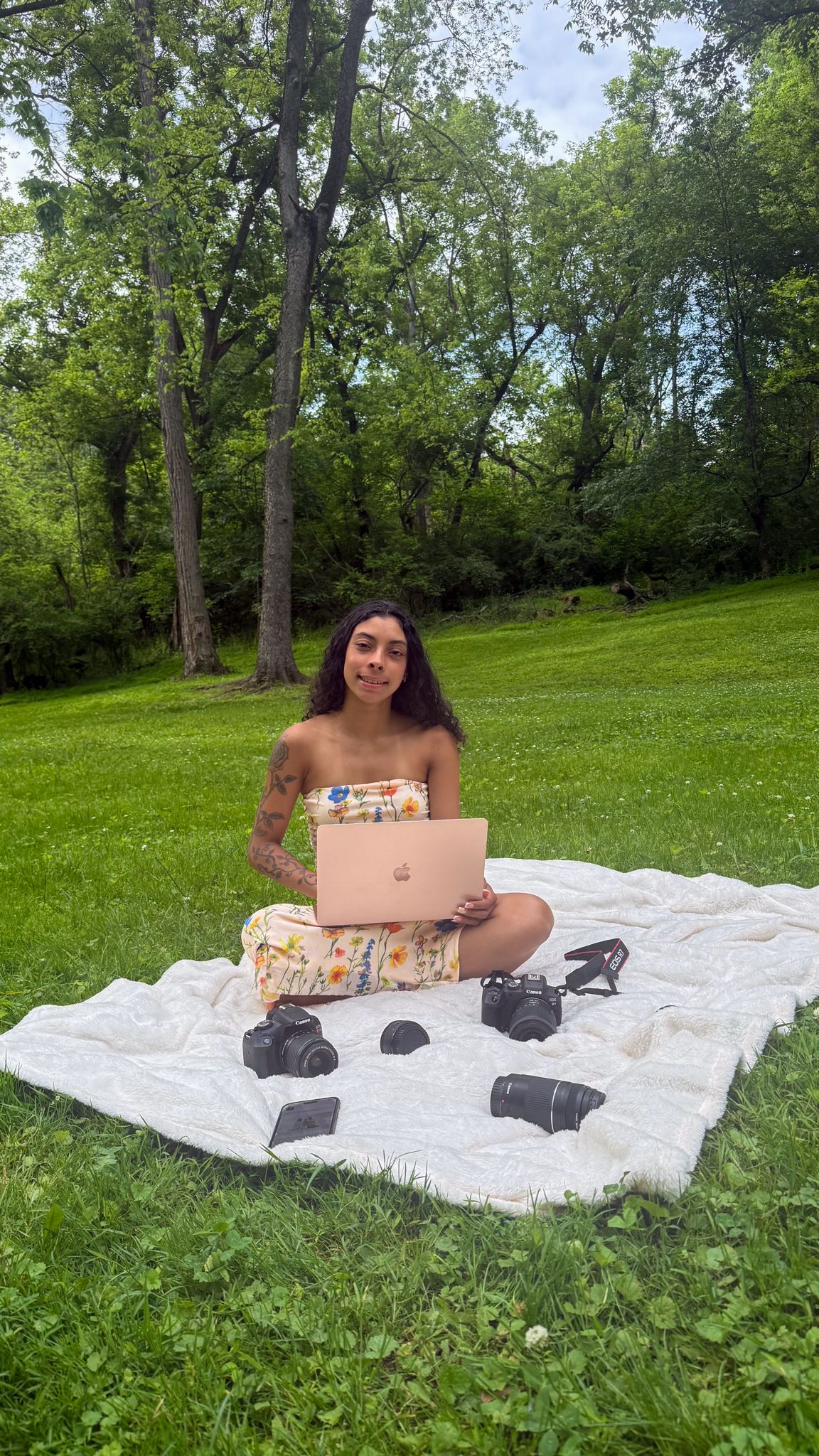 Woman sitting on blanket in park with laptop and cameras on grass surrounded by lush green trees.