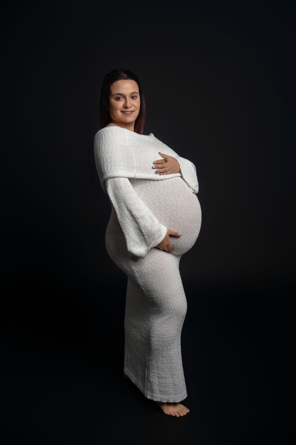 Pregnant woman in white knit dress cradling baby bump, posing for maternity photo against dark background.