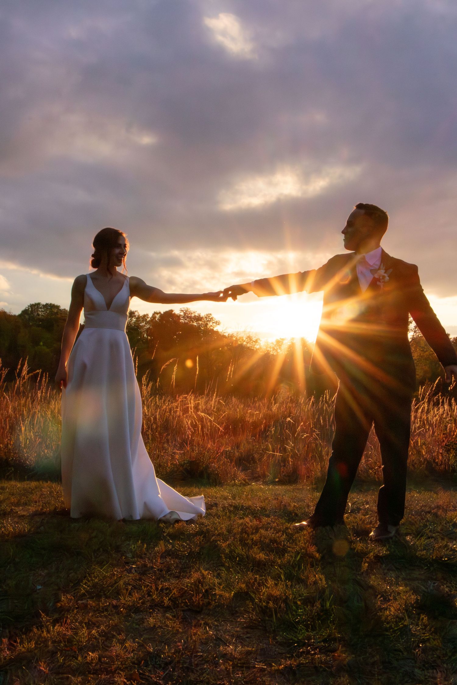 Bride and groom holding hands at sunset in a golden field in French Park in Cincinnati, silhouetted against dramatic clouds and sun rays.