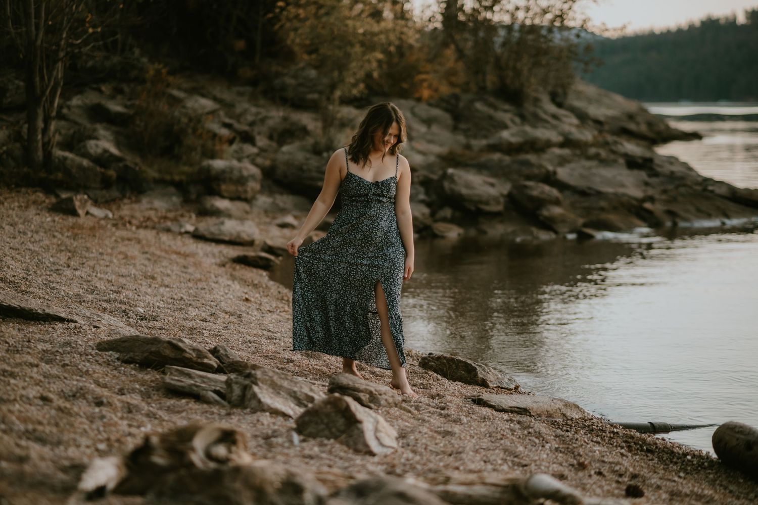 Woman in floral maxi dress walking on rocky riverbank at golden hour.