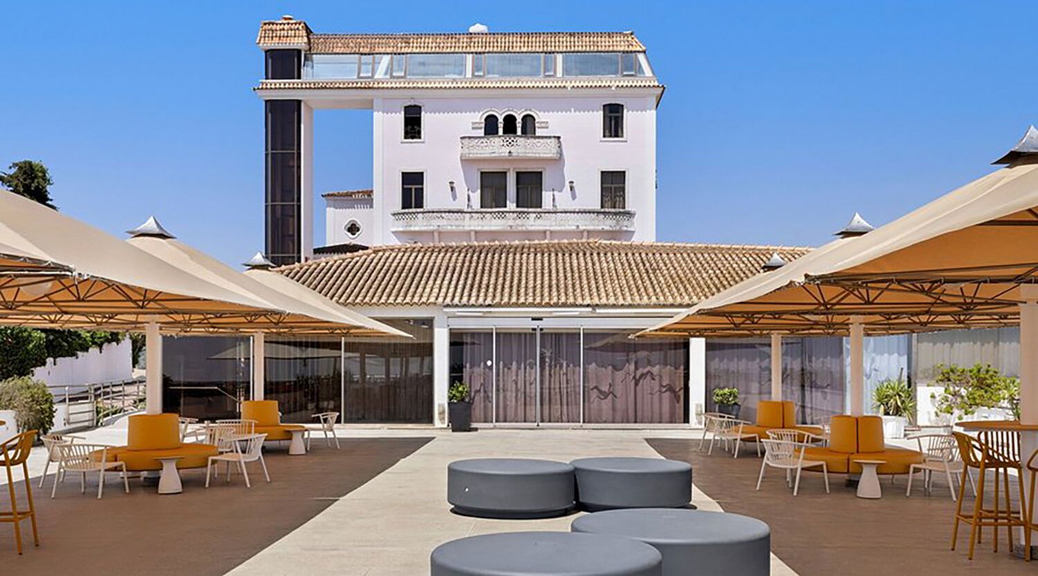 Elegant hotel terrace with beige umbrellas, colorful chairs, and a white multi-story building backdrop under clear skies.