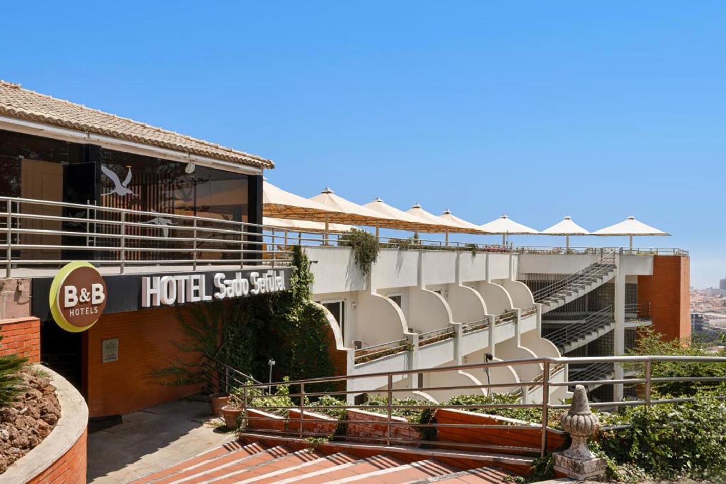 Exterior view of B&B Hotel Sainte-Tertre with terrace, umbrellas, and blue sky in background.