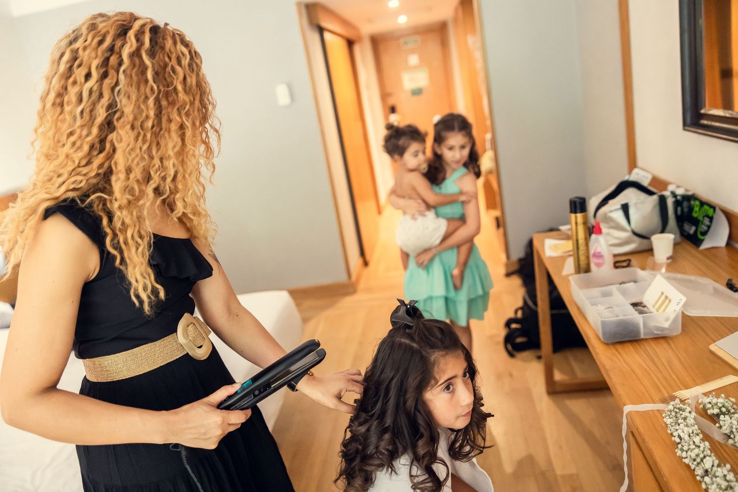Woman styling young girl's hair with flat iron in hotel room while another child is held nearby.
