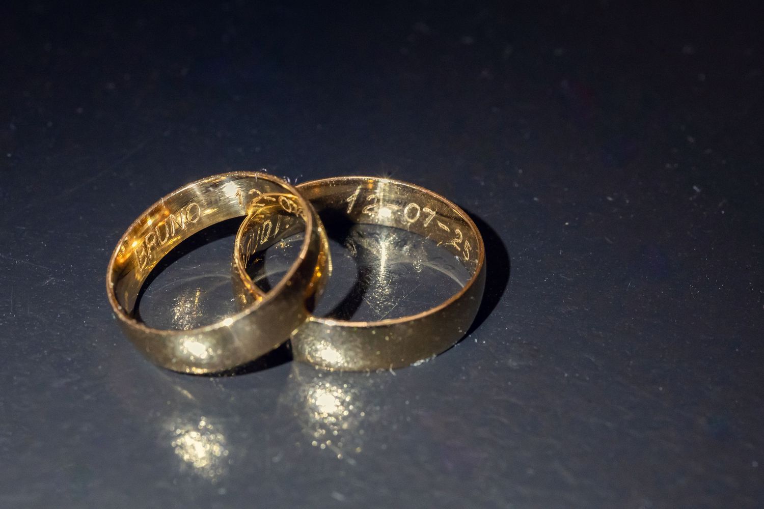 Two gold wedding bands with engraved dates resting together on a dark reflective surface.