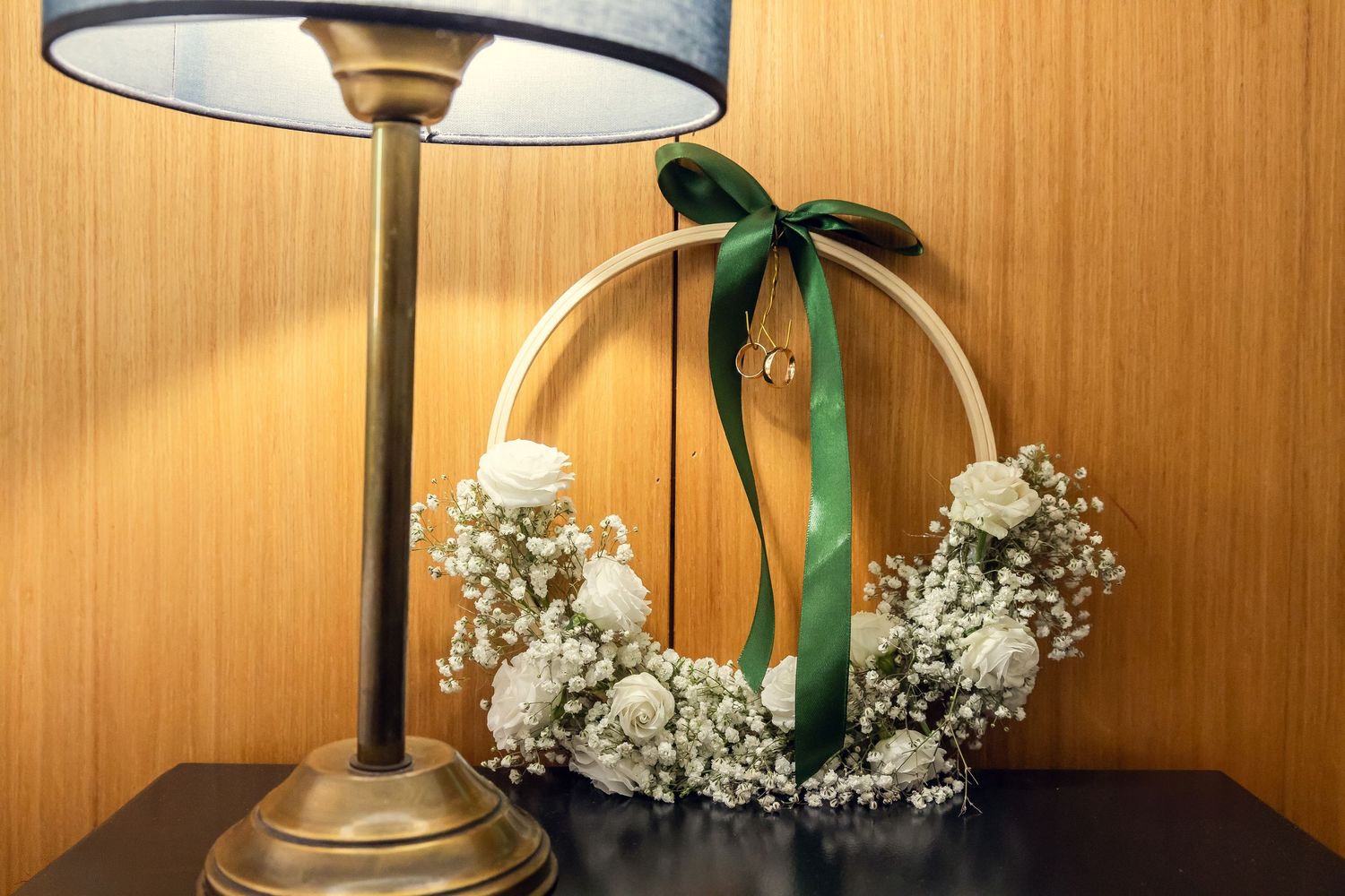 Elegant floral hoop wreath with white roses and baby's breath, adorned with a green ribbon bow on a dark table.