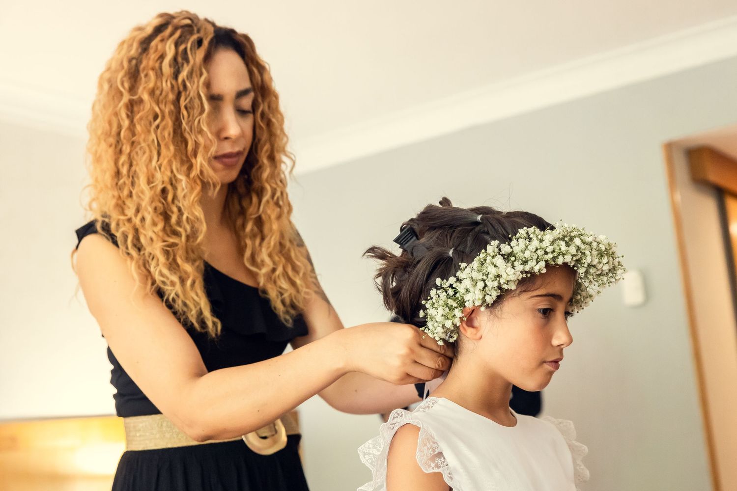 Woman with curly hair placing a floral crown on a young girl in a white dress for a wedding.