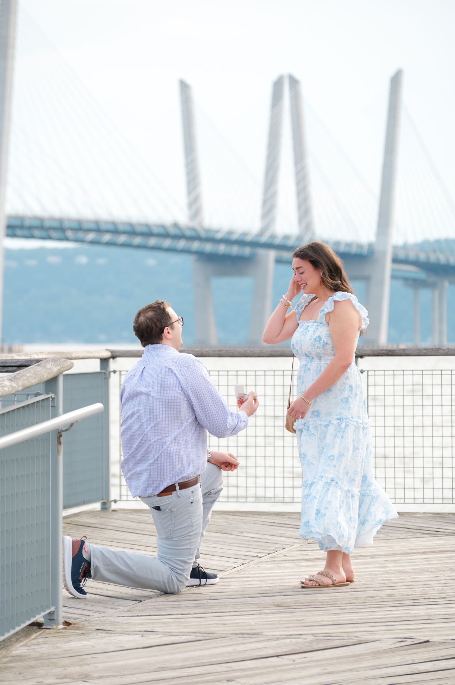 Man proposes on to woman on wooden pier at Pierson Park in Tarrytown, NY along the Hudson River with a bridge behind them.