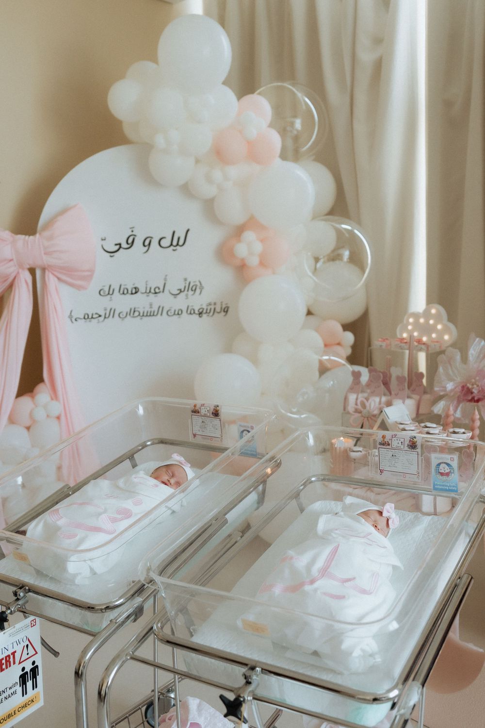 Twin newborn celebration setup with two decorated bassinets and a soft balloon arch
