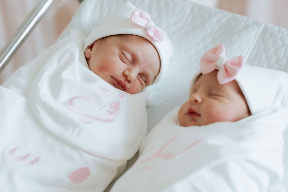 Newborn twins sleeping side by side in white swaddles and pink-bow hats