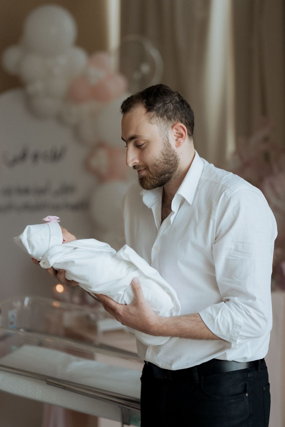 Father in a white shirt holding and gazing at his swaddled newborn amid balloon decor