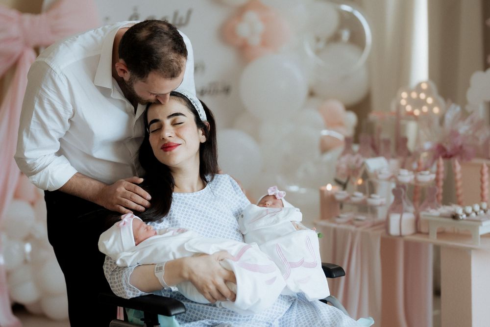 Parents in white embracing while holding their twin newborns beneath an elegant balloon arch