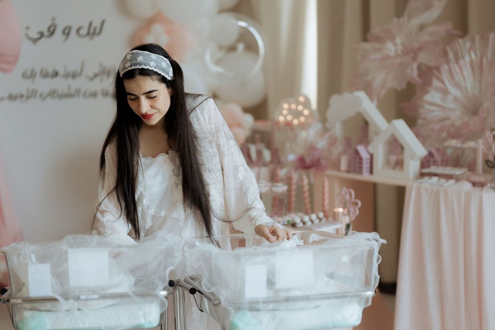 Mother in white tending to twin newborns in bassinets at a pink and white baby celebration