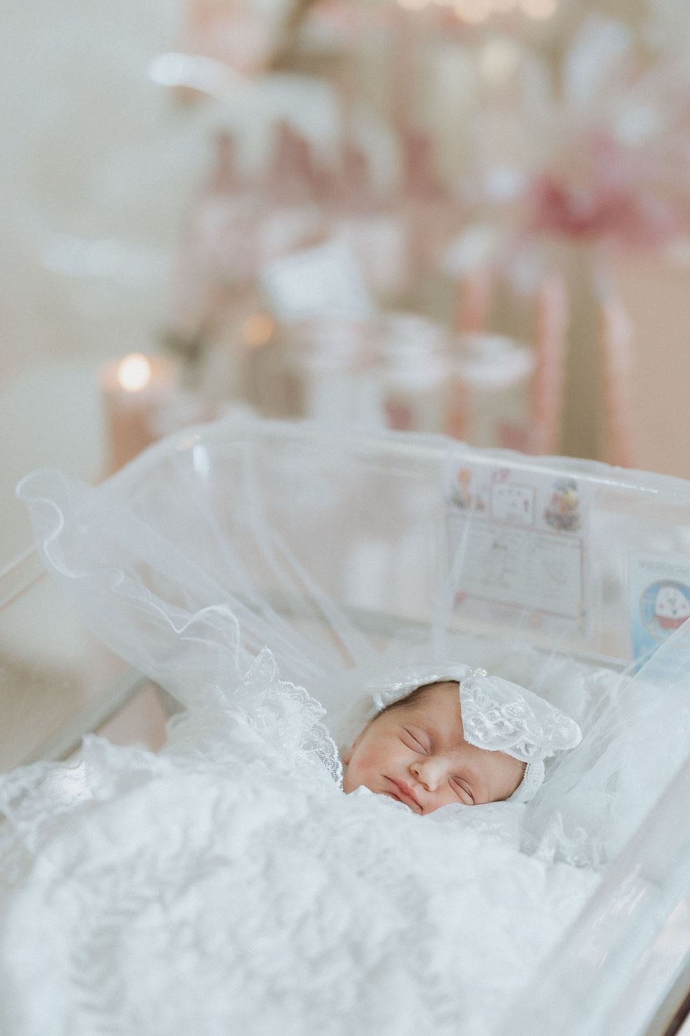 Sleeping newborn baby wearing a headband in a lace-lined bassinet with soft pink decor