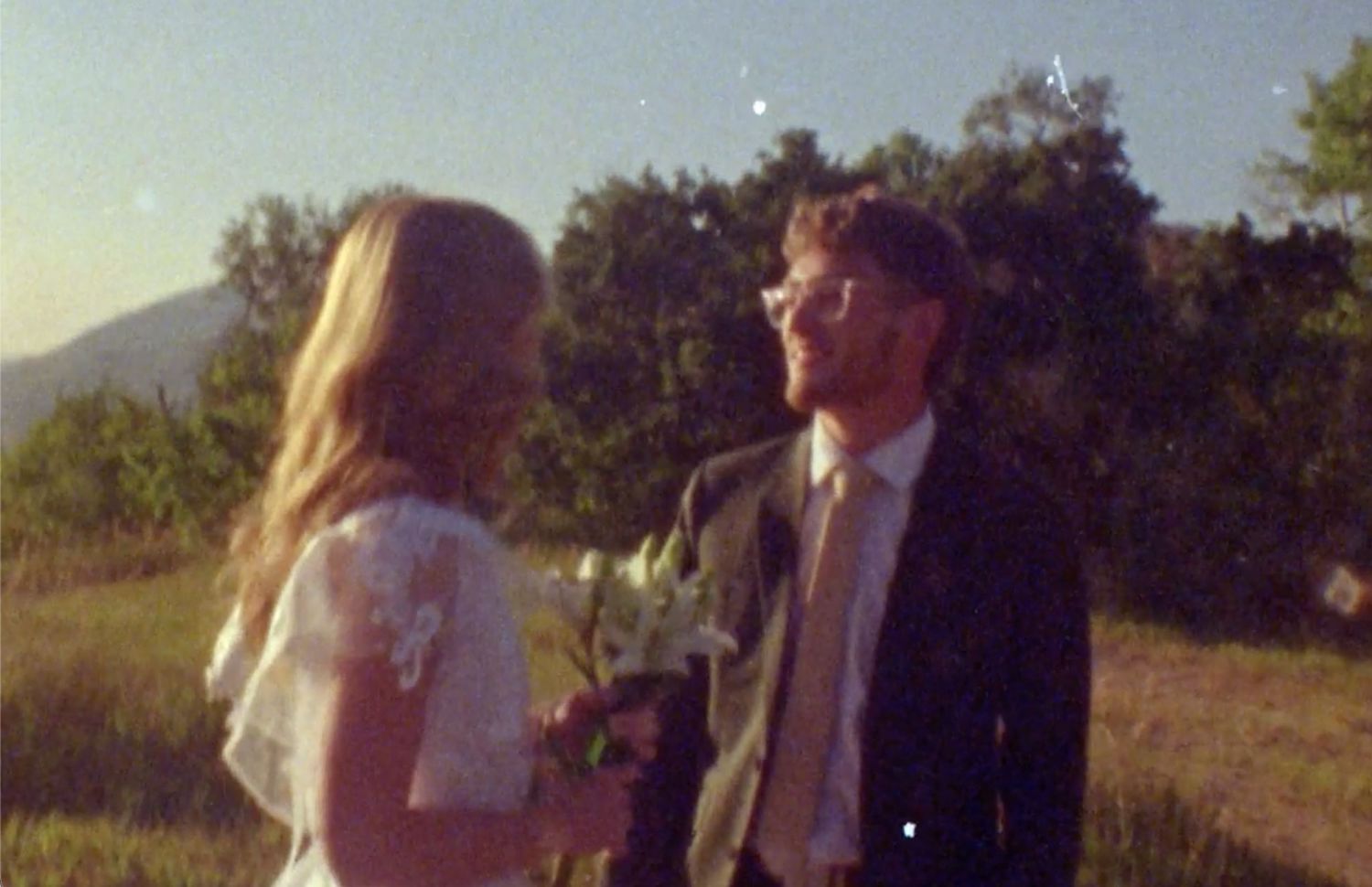 A bride in white lace and groom in suit share a romantic outdoor moment with mountains and trees in background.
