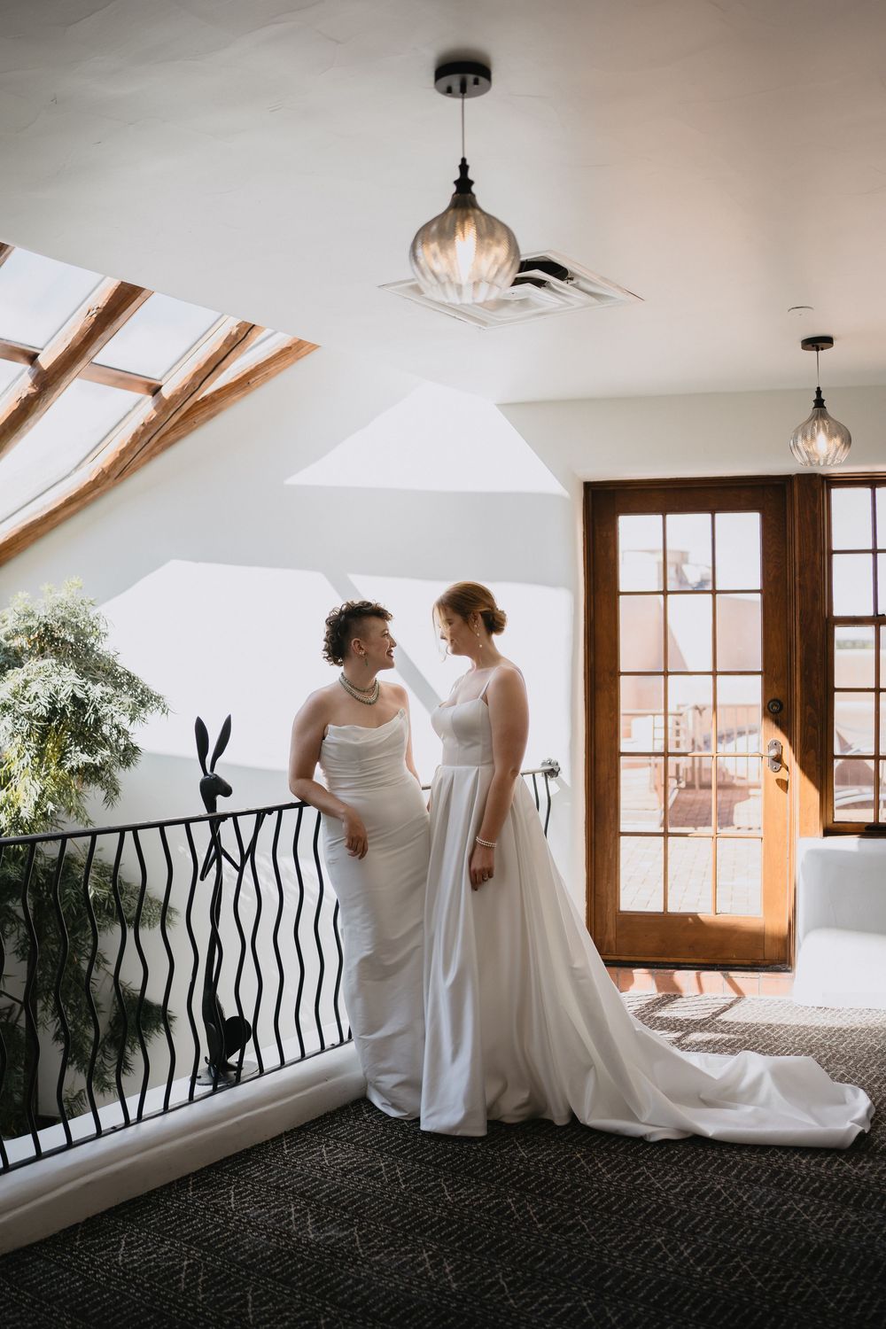 Two brides in white wedding gowns share a tender moment on an elegant indoor balcony with skylights and glass doors.