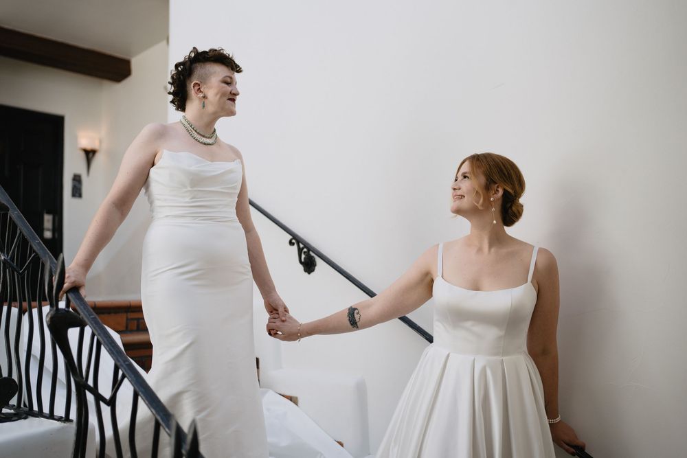 Two brides in white gowns hold hands on an elegant staircase, sharing a joyful glance on their wedding day.
