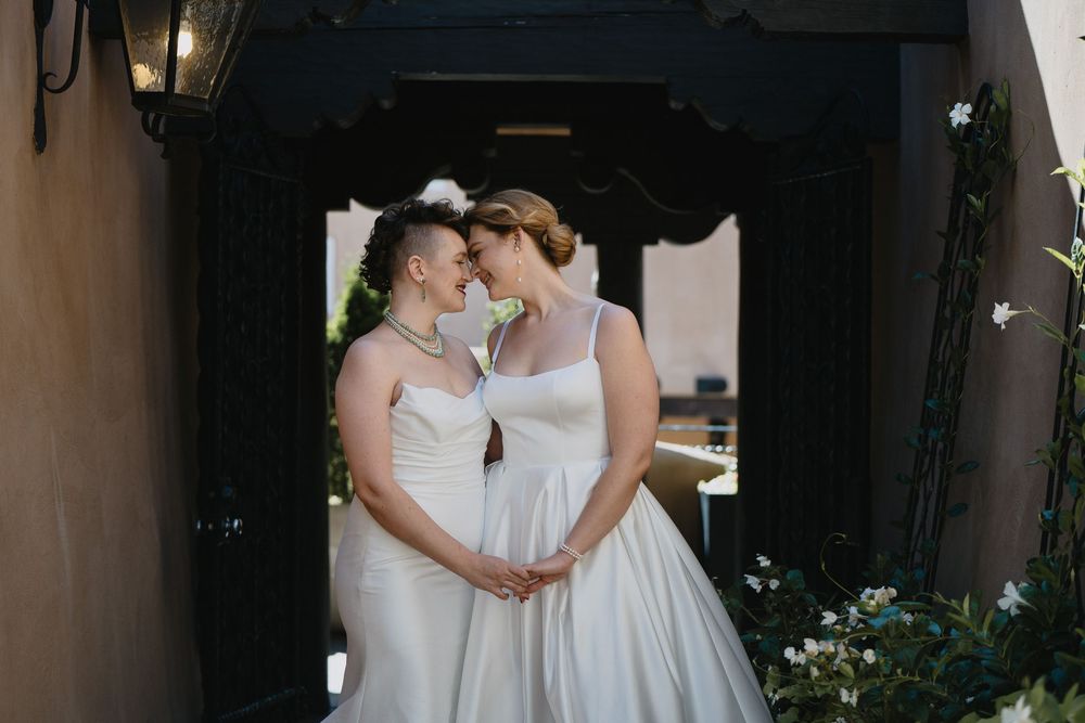 Two brides in white gowns share an intimate moment, touching foreheads while holding hands at an elegant venue.