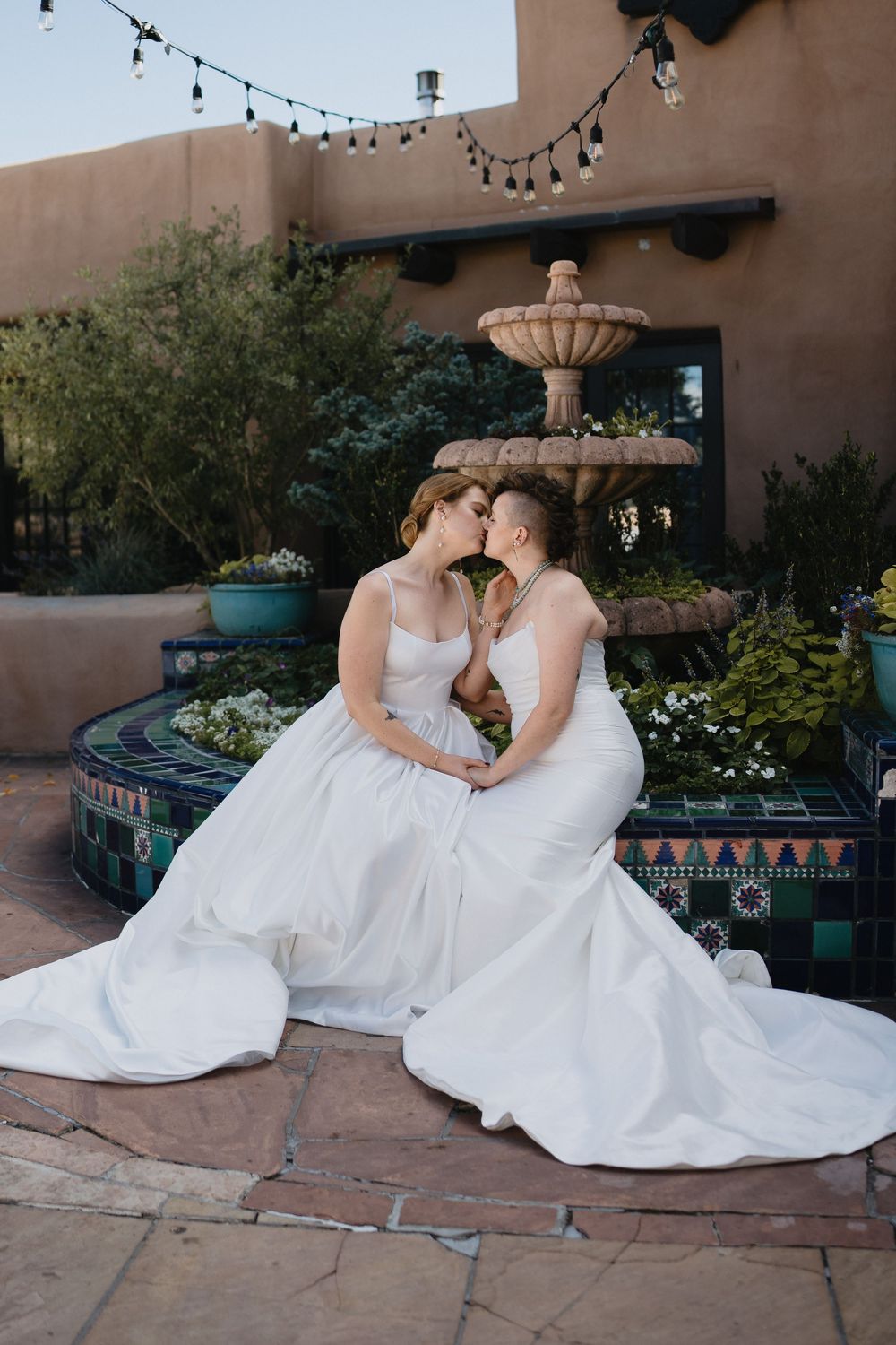 Two brides in white gowns share a kiss in front of a tiled fountain at a Southwest-style wedding venue.