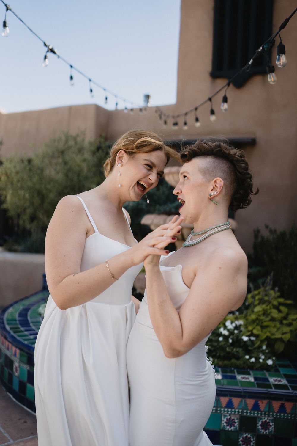Two brides in white dresses laughing and sharing cake at an outdoor wedding reception with string lights.