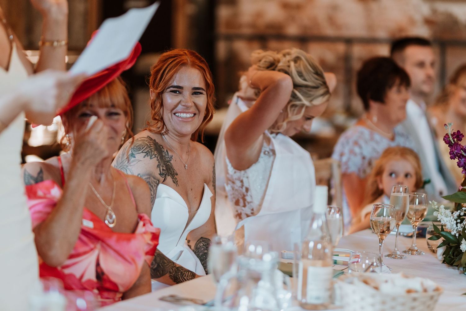 Bride with red hair and tattoos laughing joyfully at wedding reception dinner table with guests.