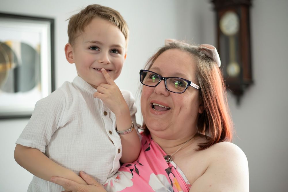 A woman with glasses and red hair holds a young boy in a white shirt, both smiling indoors near a clock.