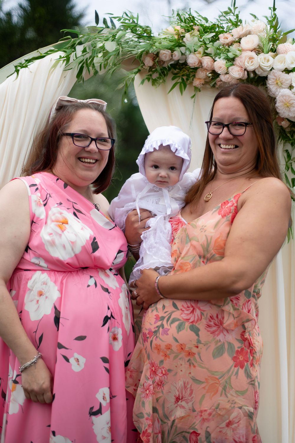 Two smiling women in floral dresses hold a baby in white christening gown under a floral arch outdoors.