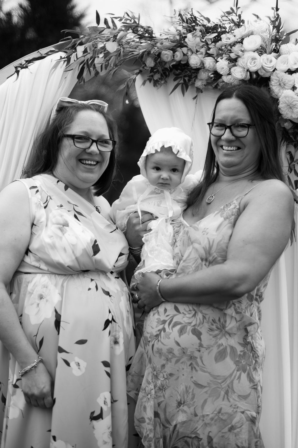 Black and white photo of two smiling women in floral dresses holding a baby in white bonnet under floral arch.