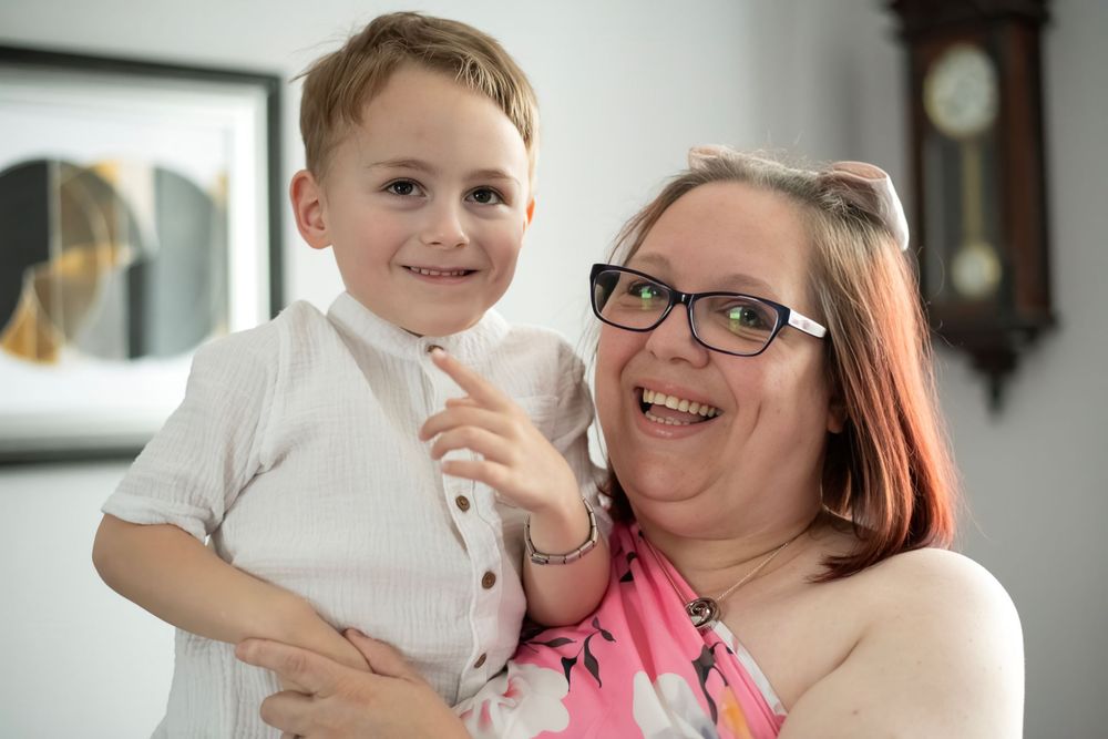 A woman with glasses and pink hair smiles while holding a young boy in a white shirt indoors.