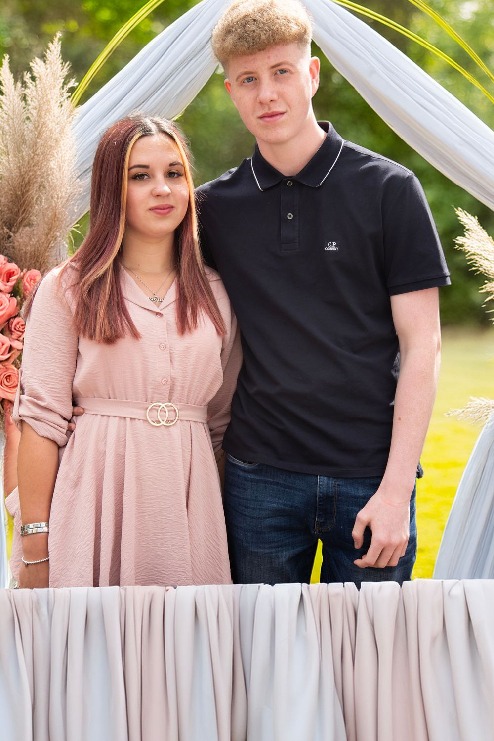 A young couple poses together under a decorated arch with flowers at an outdoor celebration.