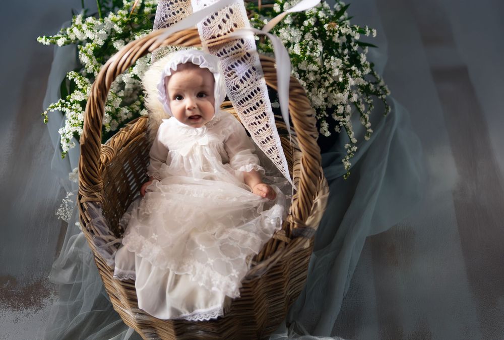 Baby in white lace christening gown and bonnet sitting in a wicker basket with white flowers.