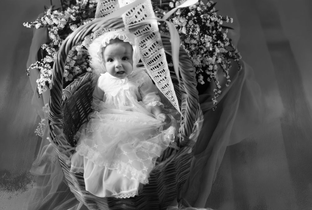 Black and white photo of a baby in white dress and bonnet sitting in a wicker basket with flowers.