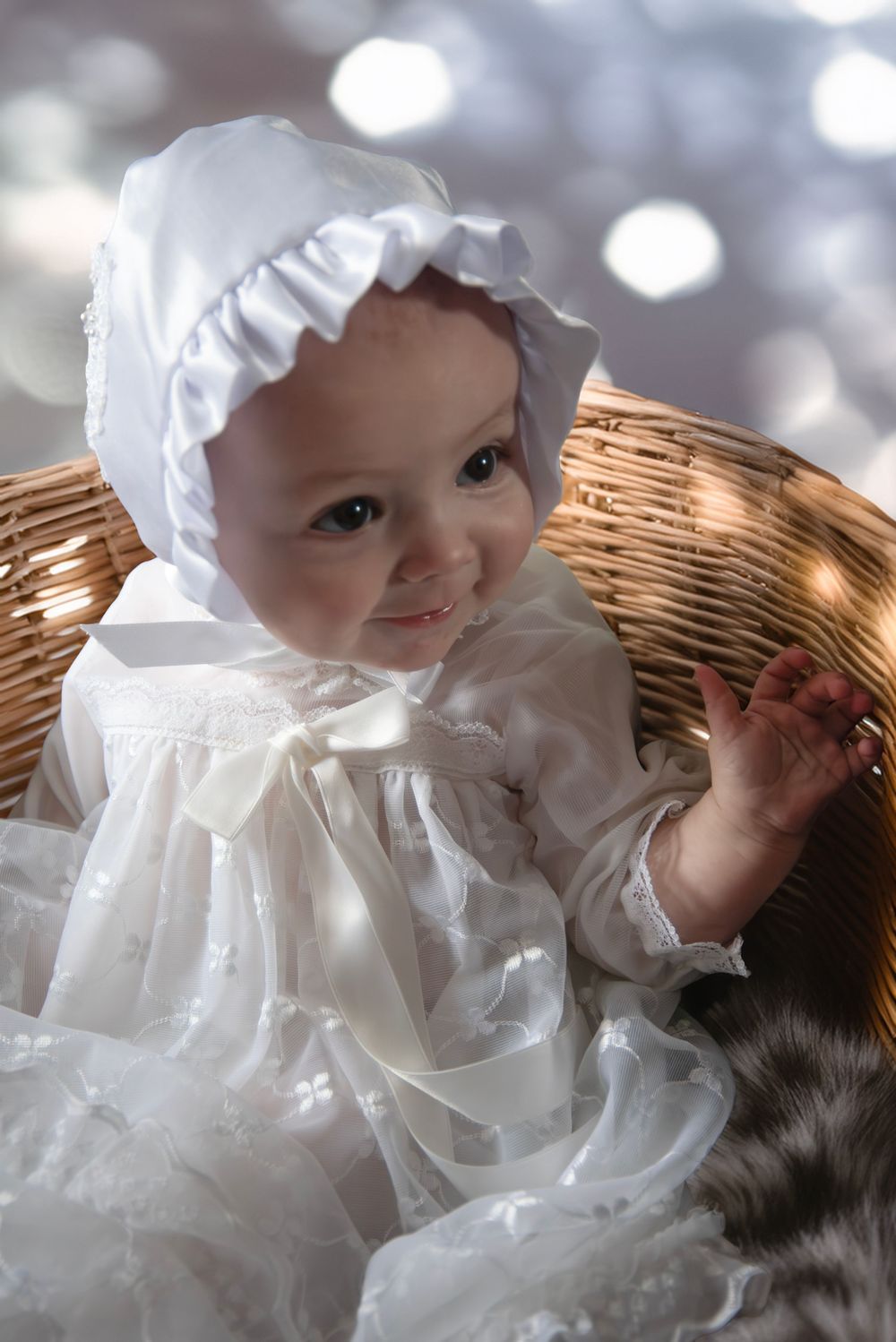Adorable baby in white christening gown and bonnet smiles while sitting in a wicker basket.