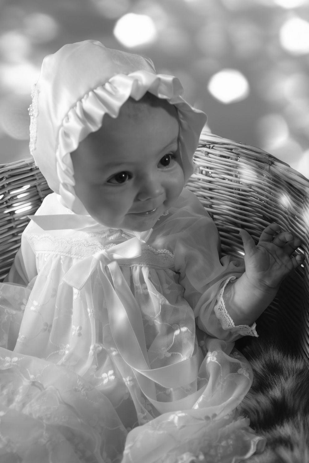 Black and white photo of a smiling baby girl in a white lace dress and bonnet sitting in a wicker chair.