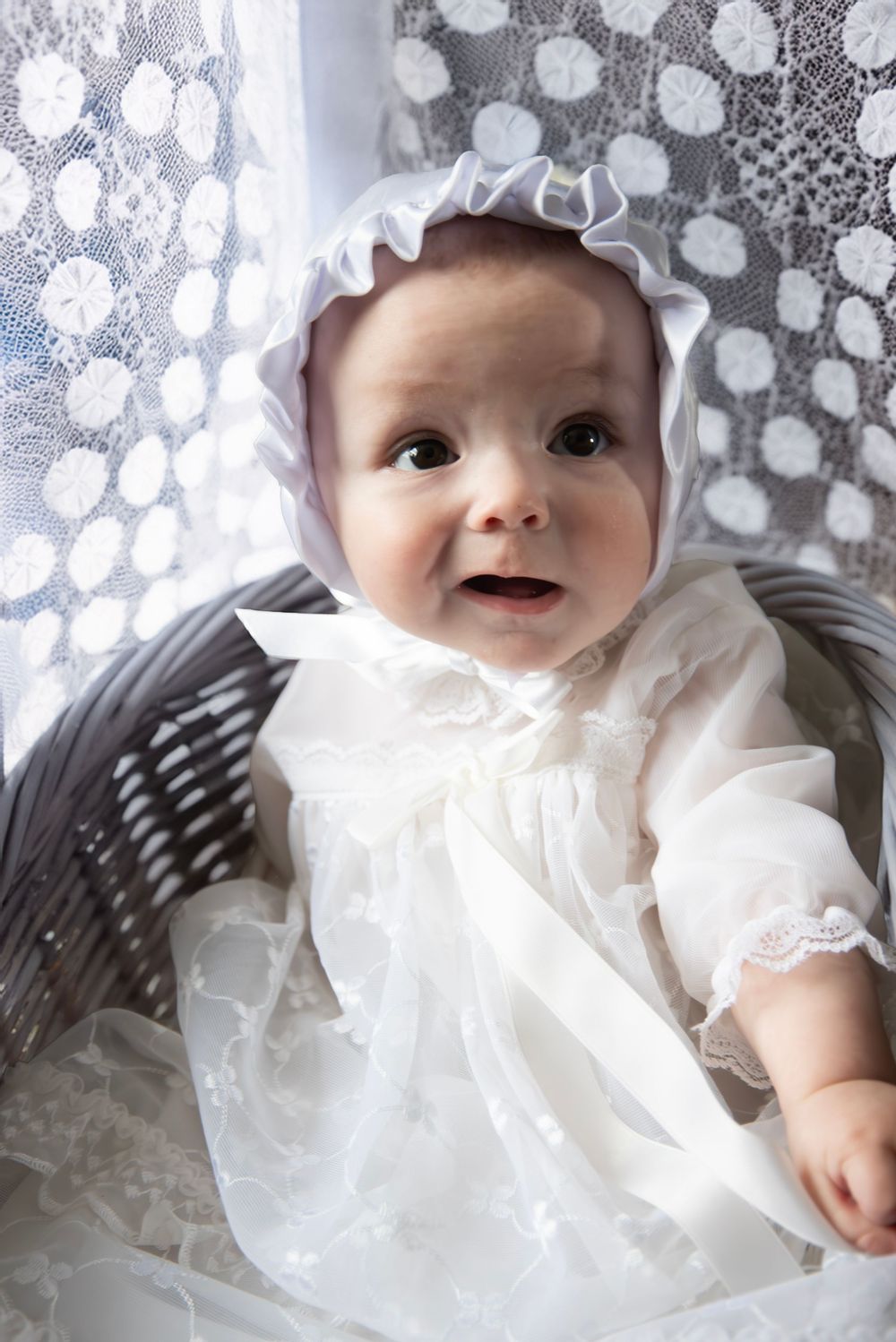 Baby in white christening gown and bonnet sitting in wicker basket near lace curtain.