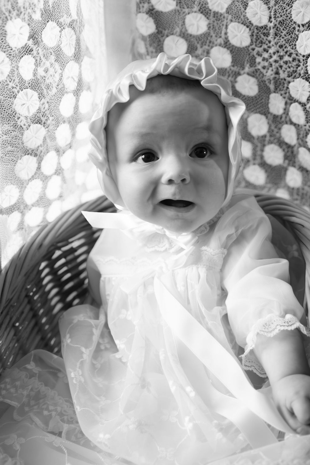 Black and white photo of a baby in a white christening gown and bonnet sitting in a wicker basket.