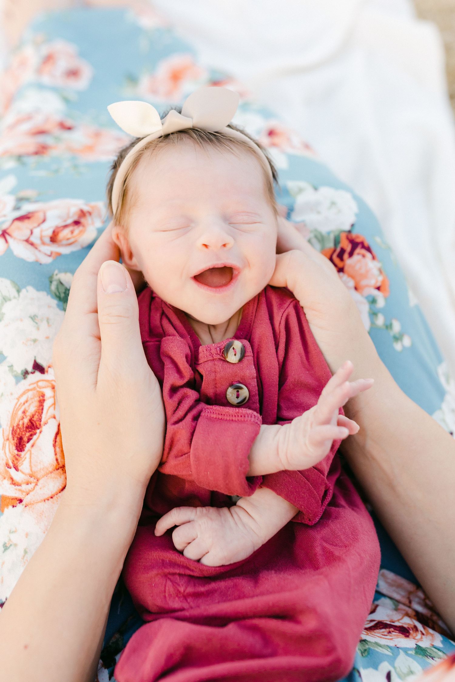 Smiling woman in floral dress holding sleeping newborn baby outdoors in golden field.