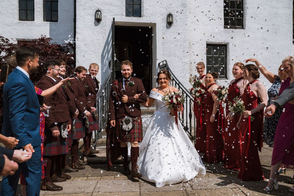 Bride and groom exit church surrounded by wedding party throwing confetti in Scottish kilts and burgundy bridesmaid dresses.