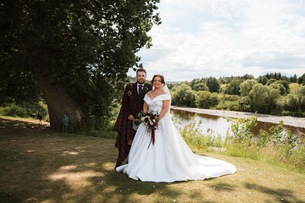Bride and groom pose by a scenic riverside with lush green trees on their wedding day in the countryside.