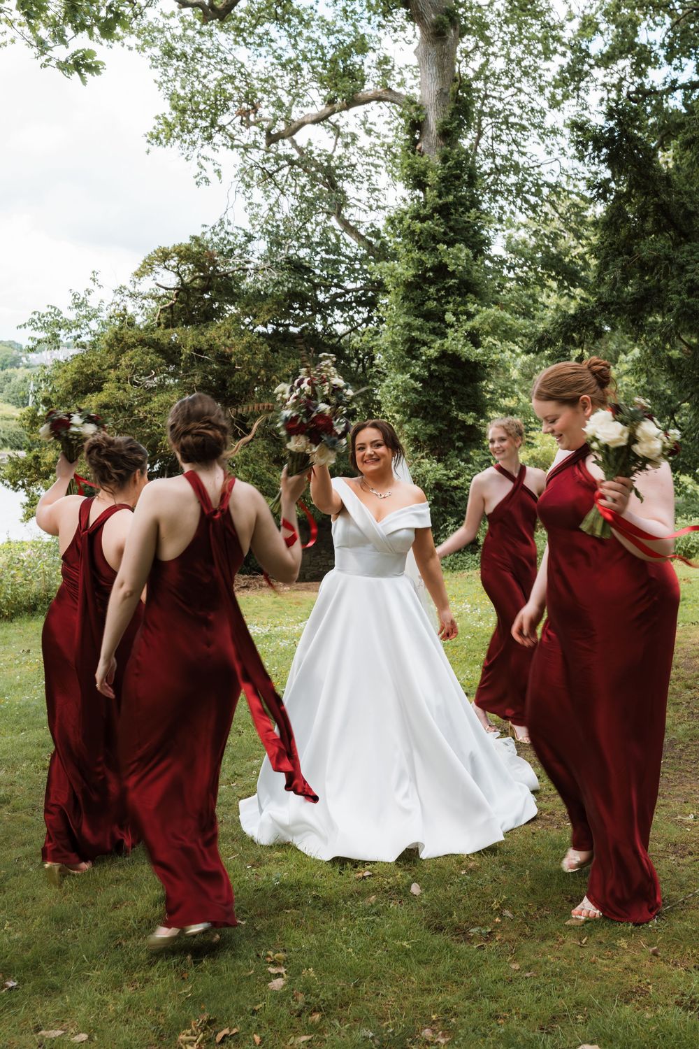 Bride in white gown surrounded by bridesmaids in red dresses dancing outdoors in lush garden setting.
