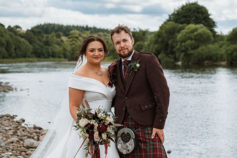 Bride and groom posing by a Scottish river, groom wearing tartan kilt and burgundy jacket, bride holding bouquet.