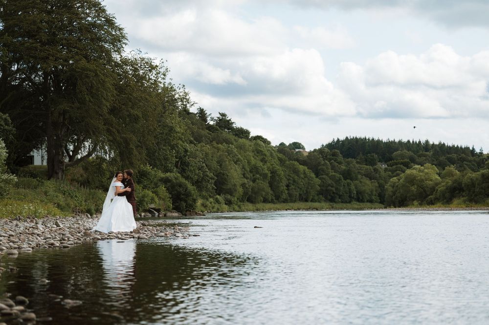 Bride and groom embracing by a scenic lakeshore surrounded by lush green trees on their wedding day.