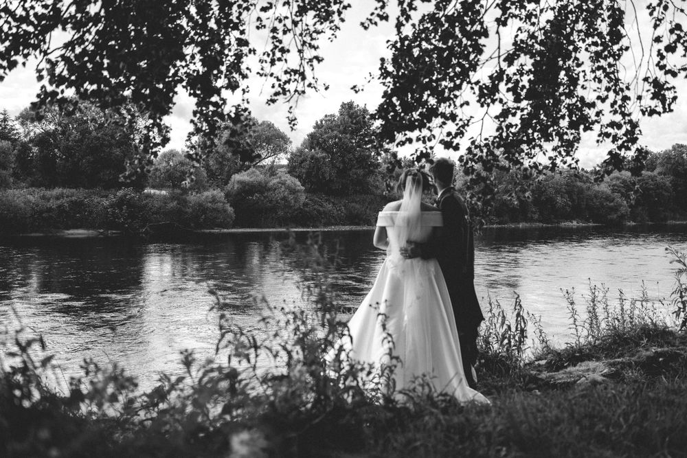 Black and white photo of a bride and groom sharing a romantic moment by a tranquil lake.